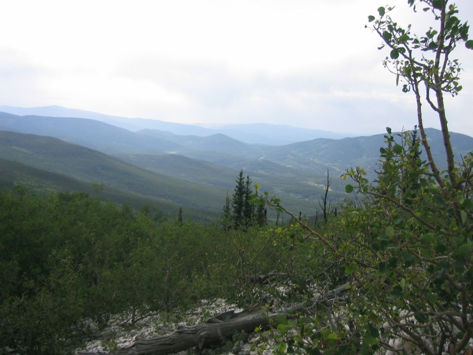 Mountain landscape with trees in the foreground and hazy blue mountains in the distance. Watermark reads SUPERMOTORS.NET