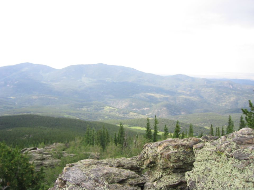 Mountain landscape with pine trees and rocks in the foreground. A watermark reads SUPERMOTORS