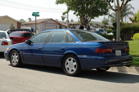 1993 Ford Taurus - A blue 1993 Ford Taurus sedan is parked on a residential street, seen from the rear quarter angle.