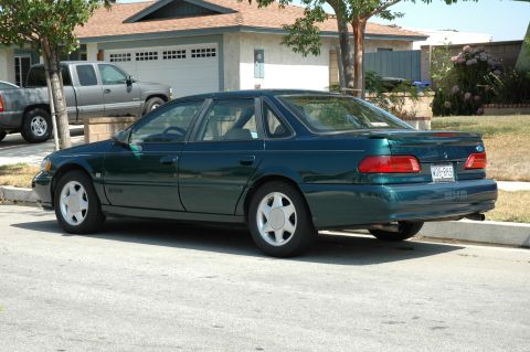 1993 Ford Taurus - A green sedan is parked on the street in front of a house, with a pickup truck behind it.