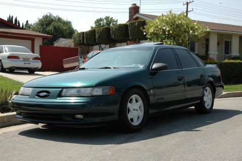 1993 Ford Taurus - A dark green 1993 Ford Taurus sedan is parked on a residential street, with another white sedan visible in the background.