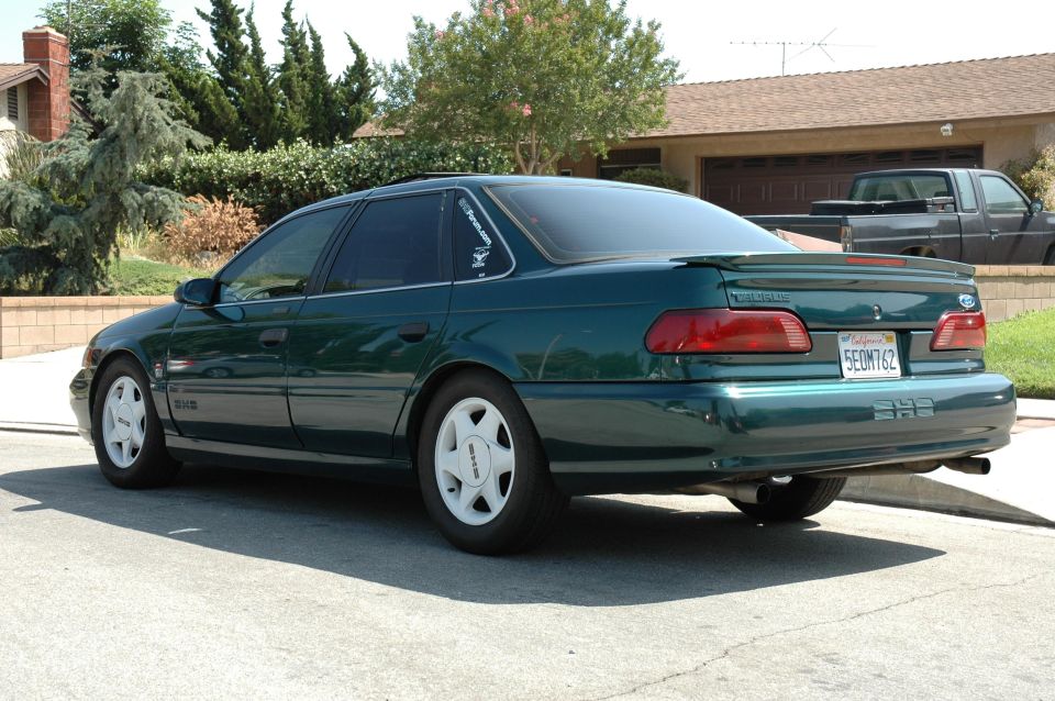 Green sedan parked on street, rear view. Another truck is parked in the driveway behind it