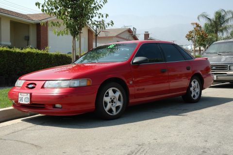 1993 Ford Taurus - A red sedan is parked on a residential street next to another vehicle.