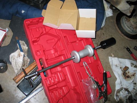 1995 Ford Bronco - A slide-hammer puller with a heavy steel slide weight laid across a red molded tool case on a garage floor, alongside snap-ring pliers, a T…