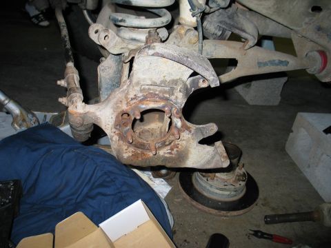 1995 Ford Bronco - Front suspension torn down on a garage floor, showing a rusty steering knuckle with the hub and rotor removed and the mounting flange and s…