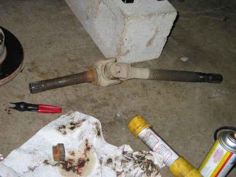 1995 Ford Bronco - Removed front axle shaft with a U-joint style joint laid on a concrete garage floor against a cinder block, showing splined ends at both si…