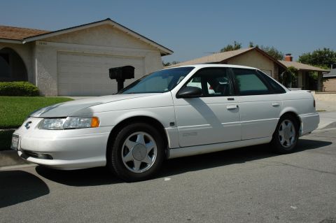 1993 Ford Taurus - White sedan parked on a street in front of a house. The hood has a silver cover.