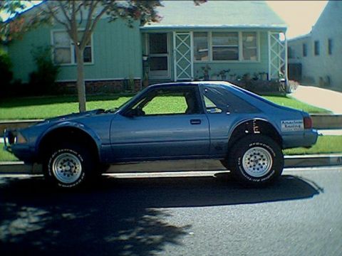 1990 Ford Bronco - lifted blue Fox-body Ford Mustang hatchback fitted with off-road tires sits at the curb of a residential street.