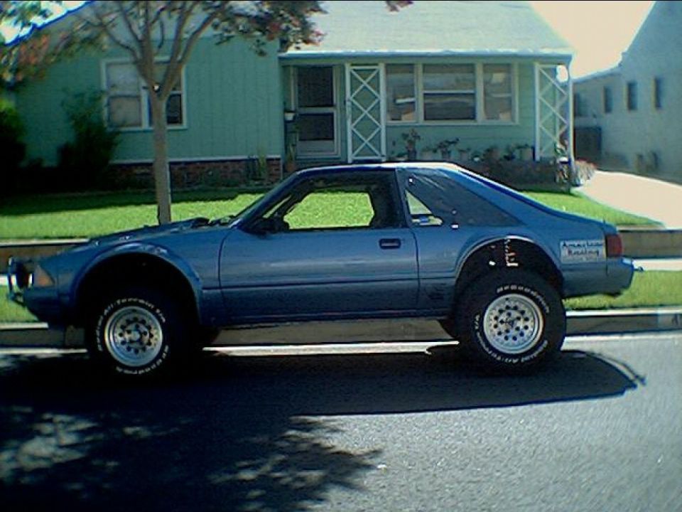 Lifted blue Fox-body Ford Mustang hatchback with off-road tires parked at a curb