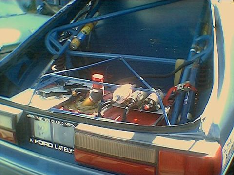 1990 Ford Bronco - View into a Ford hatchback's rear showing a blue roll cage and fuel cell, with bumper stickers reading Ford.