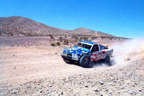 1990 Ford Bronco - off-road race truck wearing American flag stars-and-stripes livery races through the desert kicking up dust.