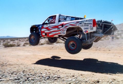 1990 Ford Bronco - Trophy truck number 803 in stars-and-stripes livery launches off a desert jump with both axles in the air.
