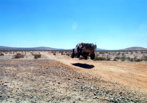 1990 Ford Bronco - off-road race truck flies through the air off a dirt berm in a wide, barren desert landscape under a blue sky.