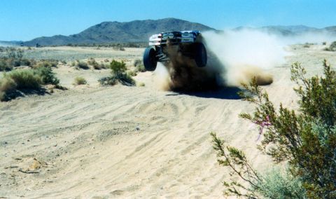 1990 Ford Bronco - off-road race truck powers through soft desert sand, throwing up a large cloud of dust near scrub brush.