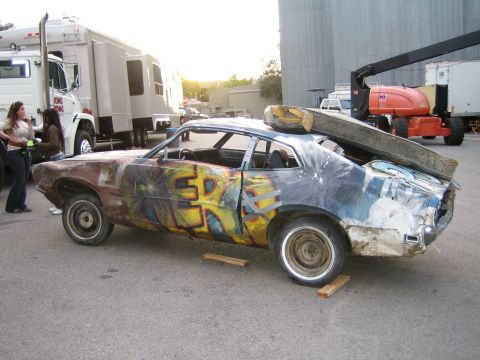 1990 Ford Bronco - rough, graffiti-painted Ford Maverick fastback rests on wood blocks in a paved lot beside RVs and equipment.