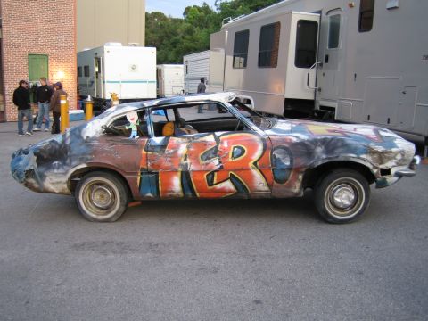 1990 Ford Bronco - Side profile of a beat-up Ford Maverick fastback covered in graffiti spelling HERO, parked behind buildings and trailers.