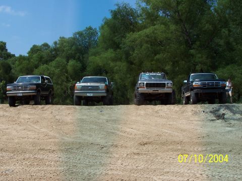 1987 Ford Bronco - A group of four trucks and SUVs are lined up on a sandy hill with trees in the background.