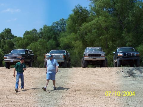 1987 Ford Bronco - Four Ford trucks are parked on a dirt hill with trees in the background.