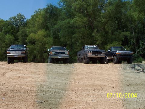 1987 Ford Bronco - A group of four pickup trucks are lined up on a sandy incline, with a dense line of trees behind them.