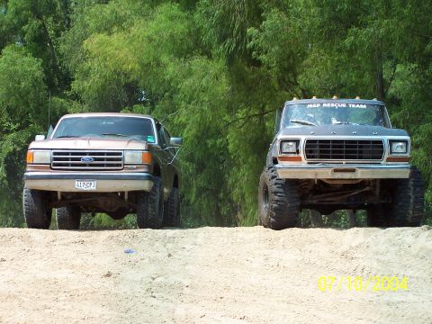 1987 Ford Bronco - Two modified Ford trucks are parked on a dirt incline with trees in the background.