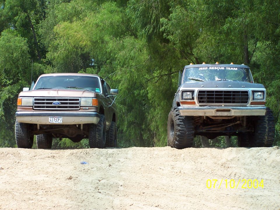 Two lifted trucks on a dirt hill, one brown Ford and one gray Ford with 'JEEP RESCUE TEAM' on top