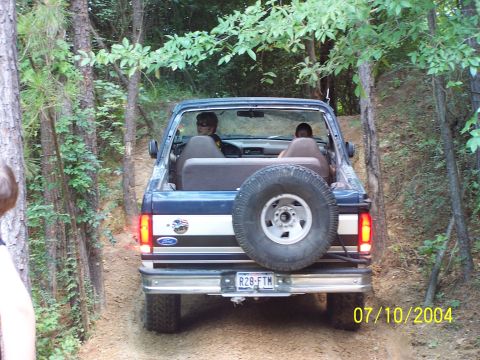 1987 Ford Bronco - This Ford Bronco is navigating a wooded trail, with two occupants visible inside the cabin.