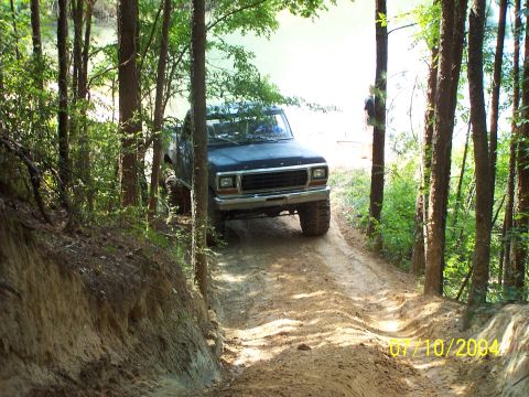 1987 Ford Bronco - A black Ford F-Series truck navigates a muddy, wooded trail with a body of water in the background.