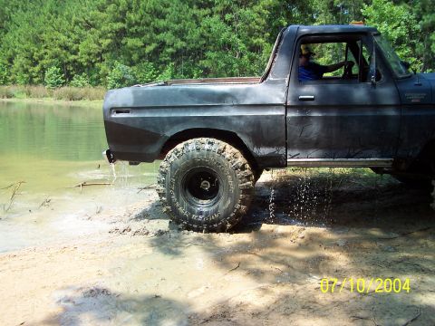 1987 Ford Bronco - A black Ford Bronco with large tires is partially submerged in water near a wooded area.