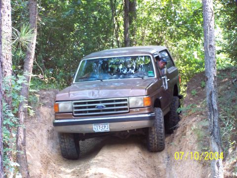 1987 Ford Bronco - A 1987 Ford Bronco navigates a challenging off-road trail surrounded by dense woods.