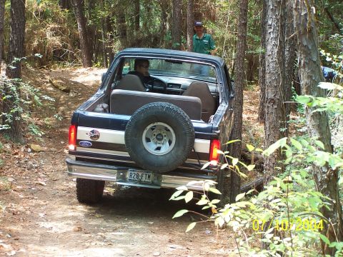 1987 Ford Bronco - A 1987 Ford Bronco navigates a wooded trail, with a spare tire mounted on the back.