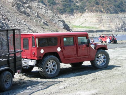1985 Ford Bronco - A red Hummer H1 wagon is parked on a dirt lot near a body of water, with other off-road vehicles visible in the distance.