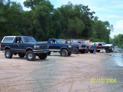 1987 Ford Bronco - Several Ford Broncos and trucks are parked on a sandy beach near trees and water.