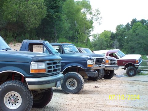 1987 Ford Bronco - Several Ford trucks are parked in a line on a dirt lot with trees in the background.