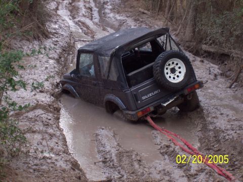 1987 Ford Bronco - A black Suzuki Samurai is deeply stuck in a muddy off-road trail, with a tow rope attached to its rear.