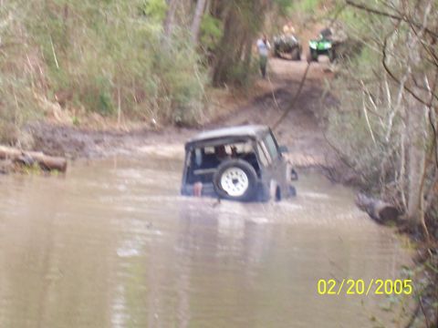 1987 Ford Bronco - Off-road vehicle navigates a muddy water crossing with other vehicles and people visible on the trail.