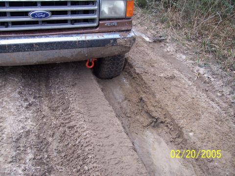 1987 Ford Bronco - A 1987 Ford Bronco navigates a muddy track, with its front tire deep in a rut and the grille visible.