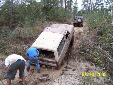 1987 Ford Bronco - Two men attempt to free a muddy 1987 Ford Bronco from a trail, with another SUV following behind.