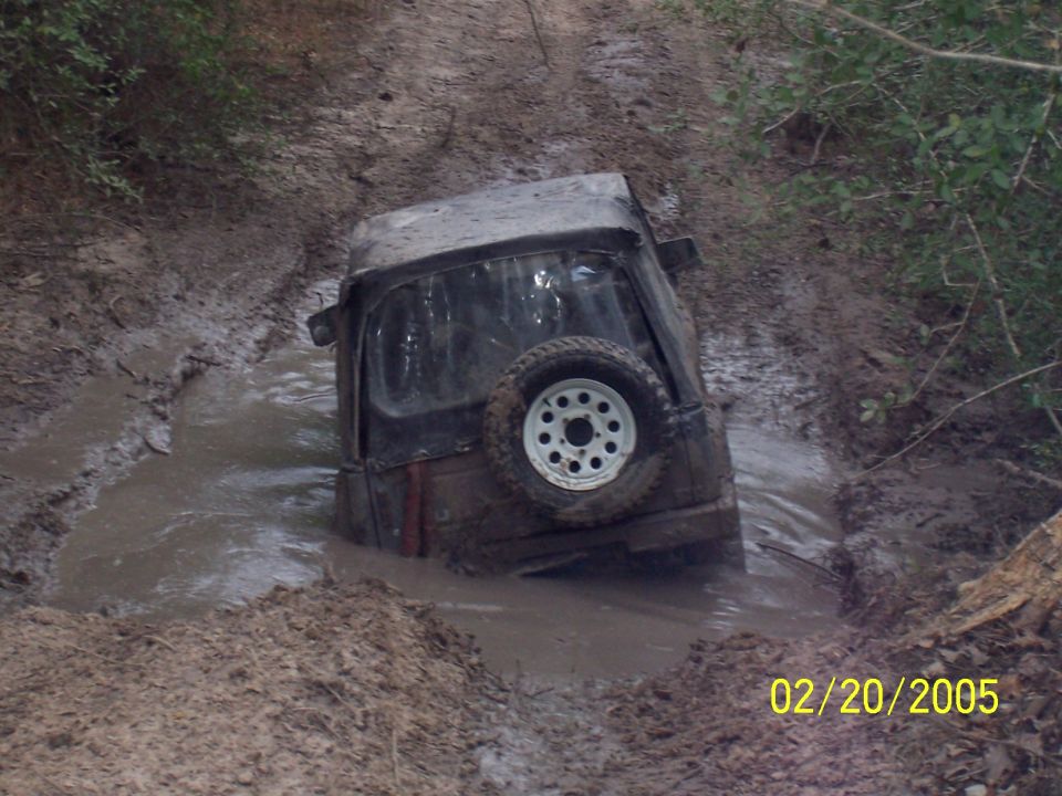 Jeep-like vehicle stuck deep in a muddy ditch, spare tire visible on the back