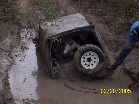 1987 Ford Bronco - An off-road vehicle is stuck deep in mud, with a person inside and another assisting.