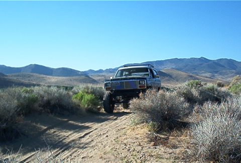1986 Ford Bronco - A lifted 1980s Ford Bronco navigates a dusty trail through a dry, mountainous landscape under a clear blue sky.