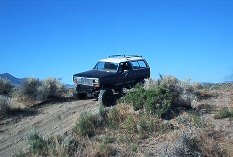 1986 Ford Bronco - A lifted Ford Bronco navigates a dusty, brush-covered incline under a clear blue sky.