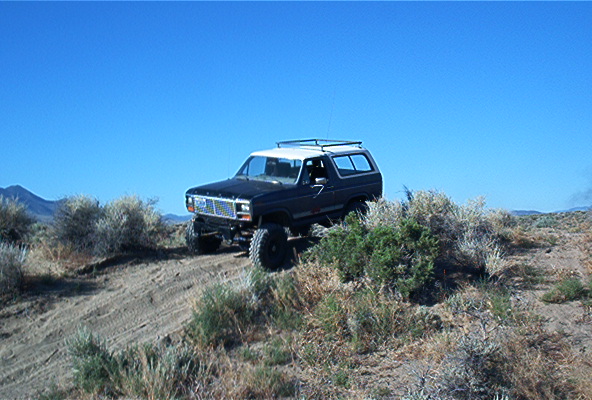 Black Ford Bronco with white top driving on a dirt hill with brush and blue sky