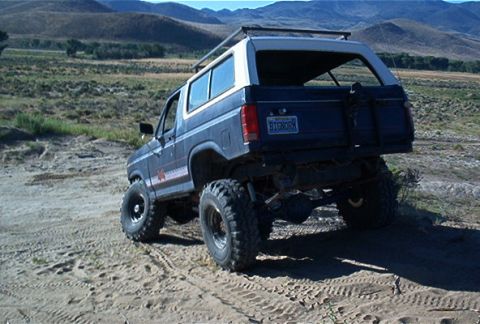 1986 Ford Bronco - This lifted Ford Bronco is navigating a dusty, off-road trail with a scenic mountain range in the distance.