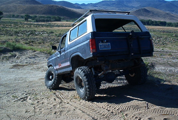Blue Ford Bronco with lifted suspension and off-road tires driving on a dirt path with mountains in the background