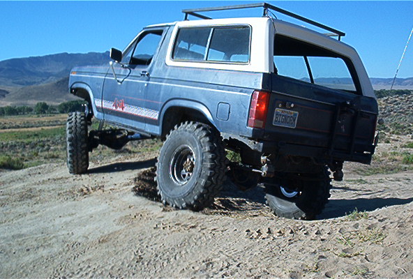 modified blue Ford Bronco with large tires on a dirt hill, white roof rack