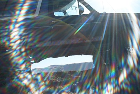 1986 Ford Bronco - Sunlight creates rainbow flares across the side of a dark SUV, with a mountainous landscape visible through the open door.