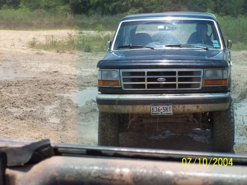 1987 Ford Bronco - A dark blue Ford Bronco navigates a muddy terrain, showing its front grille and headlights.