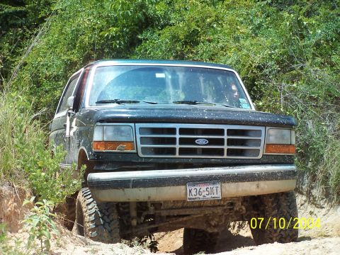 1987 Ford Bronco - A dark green Ford Bronco navigates an off-road trail with dense foliage on either side.