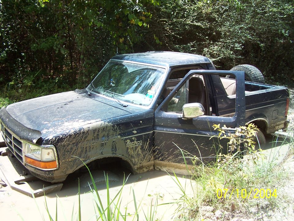 Mud-covered black Ford Bronco stuck in mud, driver's door open, spare tire visible in the bed