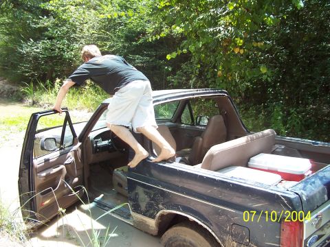 1987 Ford Bronco - Man climbs into a muddy 1987 Ford Bronco, driver's door open. Coolers visible in the rear.